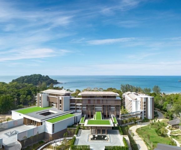 Modern beachfront resort surrounded by greenery with green rooftop terraces and ocean views under a blue sky.