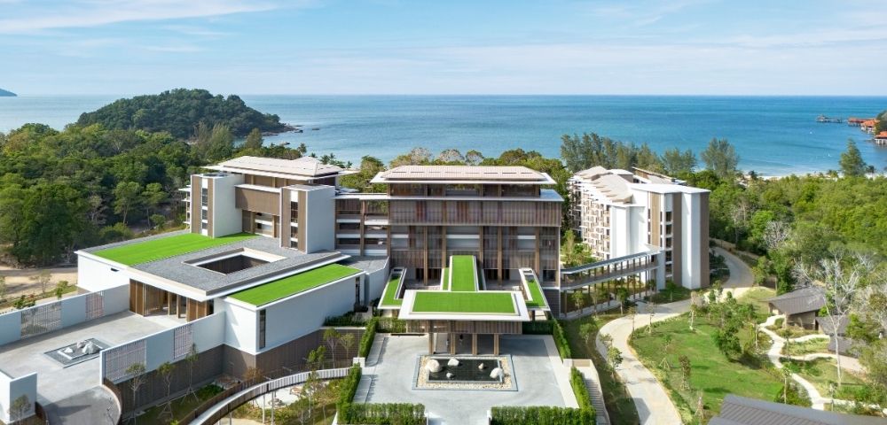 Modern beachfront resort surrounded by greenery with green rooftop terraces and ocean views under a blue sky.