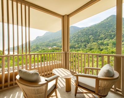 Balcony with two cushioned chairs and a small table overlooking lush green mountains under a bright sky.