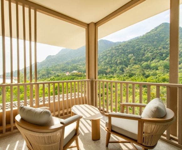 Balcony with two cushioned chairs and a small table overlooking lush green mountains under a bright sky.