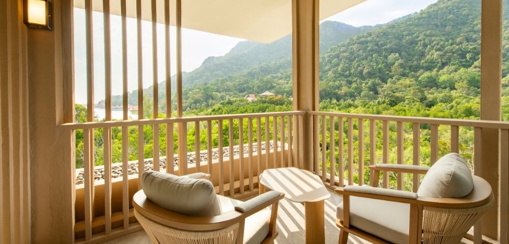 Balcony with two cushioned chairs and a small table overlooking lush green mountains under a bright sky.