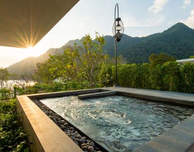 A tranquil outdoor pool with gentle ripples, nestled on a patio surrounded by lush greenery, overlooks mountains at sunset, evoking relaxation.