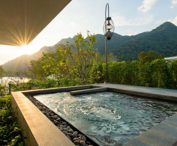 A tranquil outdoor pool with gentle ripples, nestled on a patio surrounded by lush greenery, overlooks mountains at sunset, evoking relaxation.