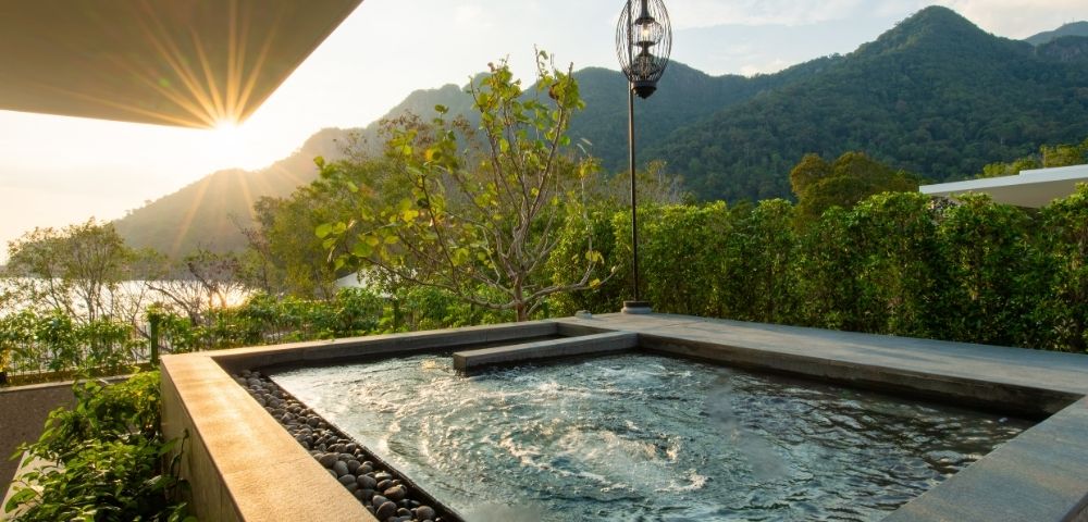 A tranquil outdoor pool with gentle ripples, nestled on a patio surrounded by lush greenery, overlooks mountains at sunset, evoking relaxation.