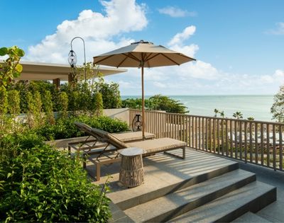 Rooftop terrace overlooking the ocean, with a lounge chair, umbrella, and lush greenery. The scene conveys tranquility and relaxation under a blue sky.