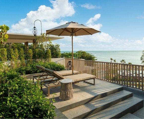 Rooftop terrace overlooking the ocean, with a lounge chair, umbrella, and lush greenery. The scene conveys tranquility and relaxation under a blue sky.