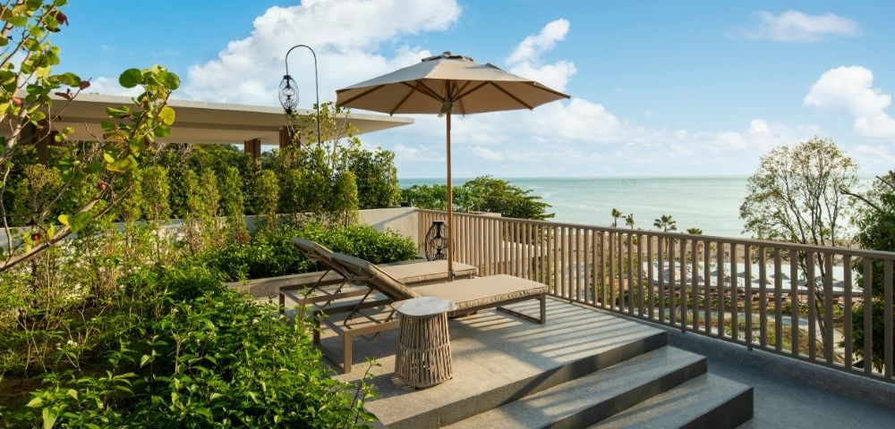 Rooftop terrace overlooking the ocean, with a lounge chair, umbrella, and lush greenery. The scene conveys tranquility and relaxation under a blue sky.