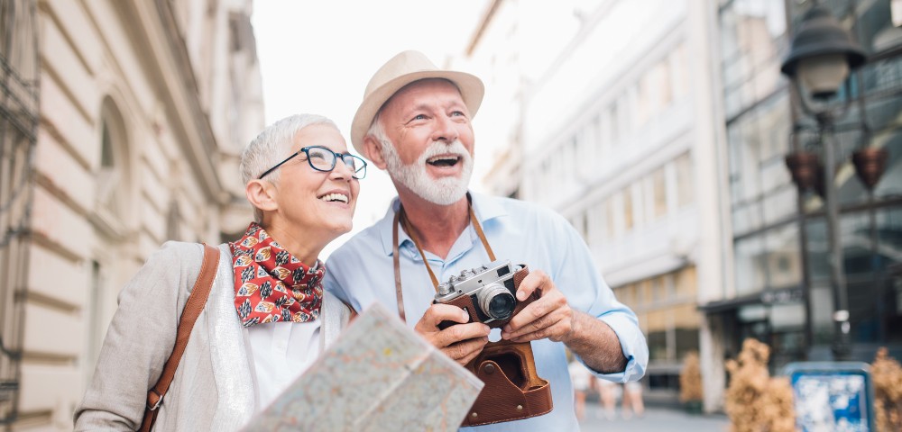 An older couple joyfully exploring a city street; the man holds a camera while the woman holds a map. They appear curious and content.