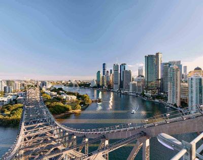 Aerial view of Brisbane cityscape with a prominent bridge crossing the river. Skyscrapers rise in the background, reflecting warm sunset light, creating a tranquil urban scene.