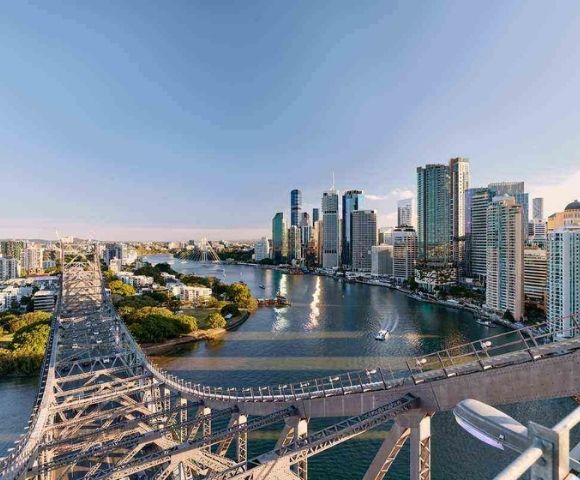 Aerial view of Brisbane cityscape with a prominent bridge crossing the river. Skyscrapers rise in the background, reflecting warm sunset light, creating a tranquil urban scene.
