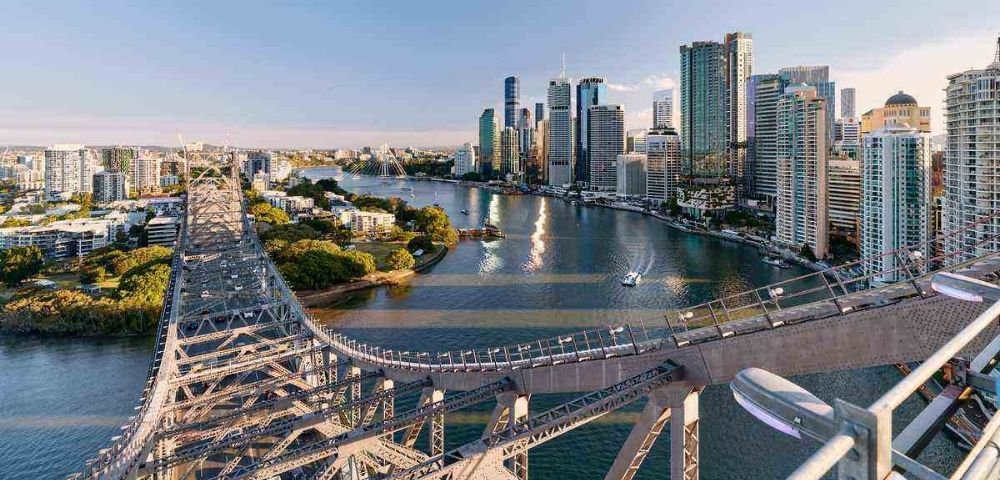 Aerial view of Brisbane cityscape with a prominent bridge crossing the river. Skyscrapers rise in the background, reflecting warm sunset light, creating a tranquil urban scene.