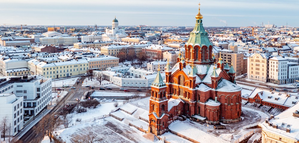 Historic red-brick cathedral with green domes stands amidst a snow-covered cityscape, featuring classical buildings under a clear, cold winter sky.
