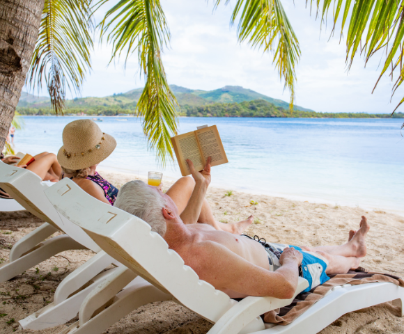 A couple relaxes on lounge chairs on a sandy beach under palm fronds. The man reads a book, and a woman in a sunhat sits beside him. Calm ocean and hills in the background convey tranquility.