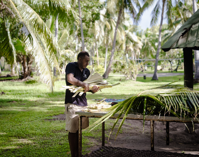 Person arranging palm leaves on a table in a tropical outdoor setting with palm trees and green grass.