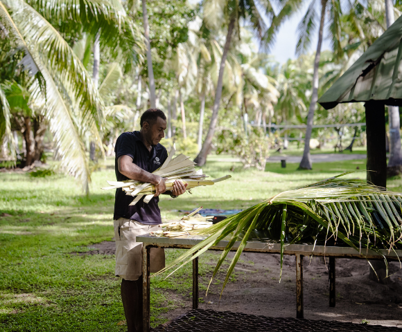 Person arranging palm leaves on a table in a tropical outdoor setting with palm trees and green grass.