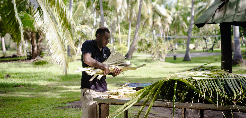 Person arranging palm leaves on a table in a tropical outdoor setting with palm trees and green grass.