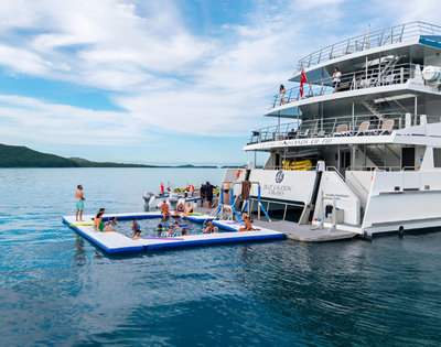 A large white yacht anchored with people swimming and relaxing on an inflatable pool platform next to it in calm blue waters.