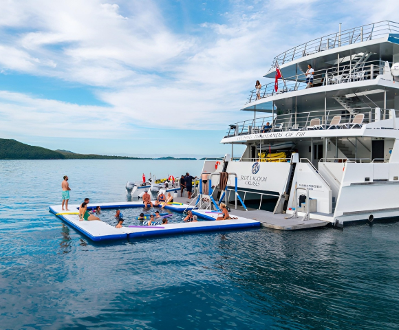 A large white yacht anchored with people swimming and relaxing on an inflatable pool platform next to it in calm blue waters.