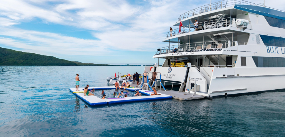 A large white yacht anchored with people swimming and relaxing on an inflatable pool platform next to it in calm blue waters.