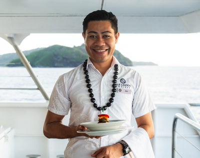 Smiling man in a white shirt holds a plate with a small burger, wearing a necklace. Background shows a scenic ocean and island view from a boat. Calm and cheerful tone.