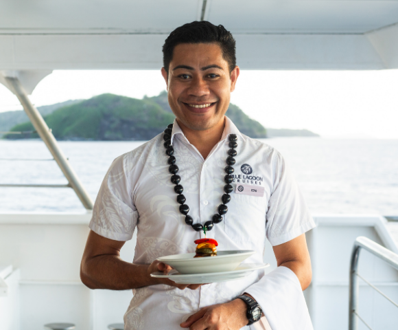 Smiling man in a white shirt holds a plate with a small burger, wearing a necklace. Background shows a scenic ocean and island view from a boat. Calm and cheerful tone.