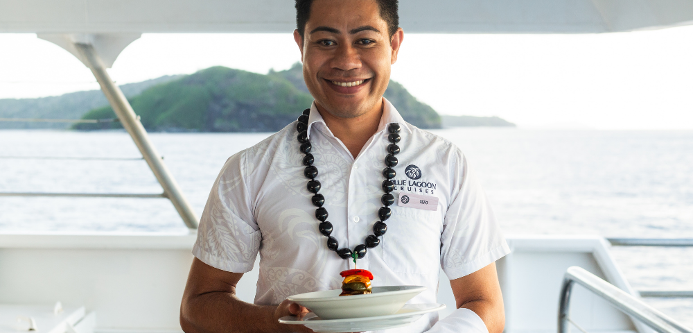 Smiling man in a white shirt holds a plate with a small burger, wearing a necklace. Background shows a scenic ocean and island view from a boat. Calm and cheerful tone.