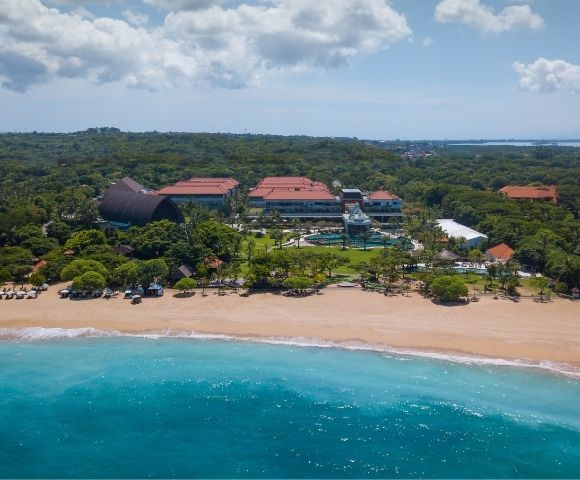 Aerial view of a beachfront resort with red-roofed buildings, swimming pool, sandy beach, and turquoise ocean under a partly cloudy sky.