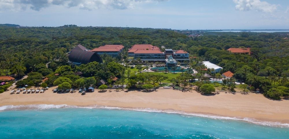 Aerial view of a beachfront resort with red-roofed buildings, swimming pool, sandy beach, and turquoise ocean under a partly cloudy sky.