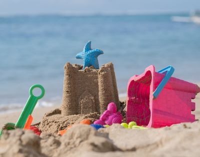 Sandcastle with a blue starfish toy on top, colorful plastic molds and a green shovel on a sandy beach by the sea.