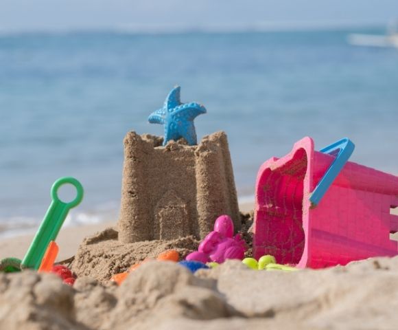 Sandcastle with a blue starfish toy on top, colorful plastic molds and a green shovel on a sandy beach by the sea.