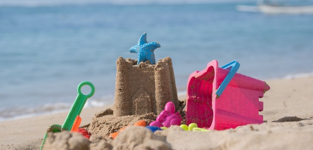 Sandcastle with a blue starfish toy on top, colorful plastic molds and a green shovel on a sandy beach by the sea.