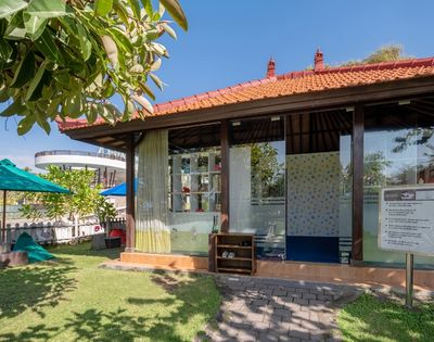 A cozy building with a red-tiled roof and glass walls in a sunny garden, surrounded by greenery and blue umbrellas, exudes a serene, inviting atmosphere.