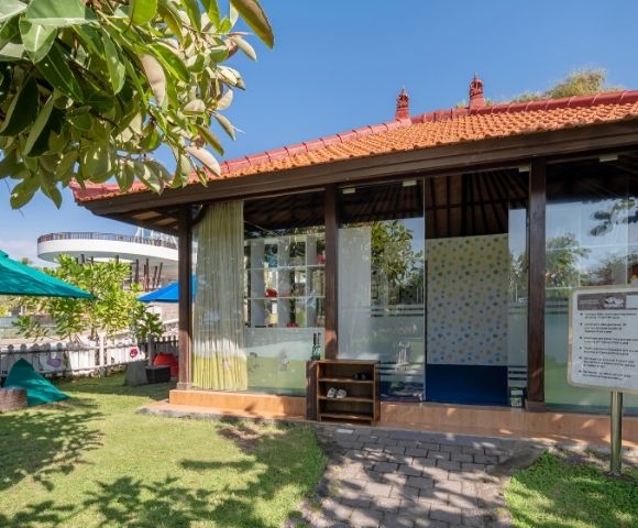 A cozy building with a red-tiled roof and glass walls in a sunny garden, surrounded by greenery and blue umbrellas, exudes a serene, inviting atmosphere.