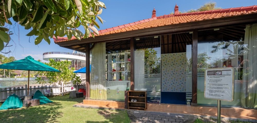 A cozy building with a red-tiled roof and glass walls in a sunny garden, surrounded by greenery and blue umbrellas, exudes a serene, inviting atmosphere.