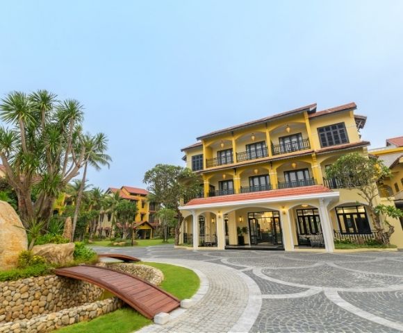 Elegant three-story yellow building with red roof tiles, framed by palm trees and lush greenery. Curved stone path and wooden bridge in foreground.