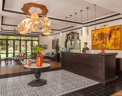 A hotel lobby with elegant lighting, ornate black and white tiles, wooden reception desk, vibrant art, and large windows overlooking a garden.