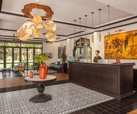 A hotel lobby with elegant lighting, ornate black and white tiles, wooden reception desk, vibrant art, and large windows overlooking a garden.