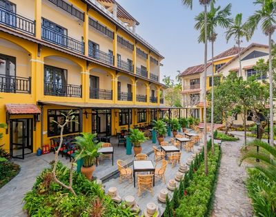 A vibrant yellow hotel with balconies overlooks a lush courtyard patio filled with wicker chairs and tables, surrounded by tropical plants and palm trees.