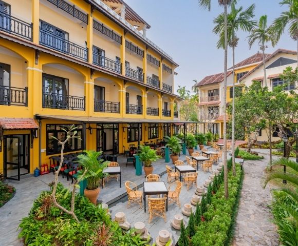 A vibrant yellow hotel with balconies overlooks a lush courtyard patio filled with wicker chairs and tables, surrounded by tropical plants and palm trees.