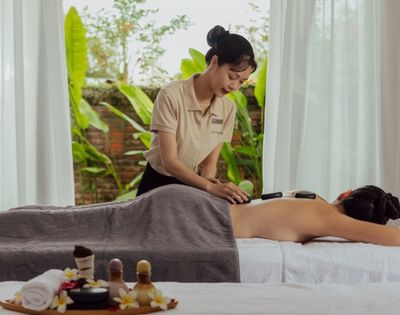 Spa therapist performs a hot stone massage on a relaxed client lying face down on a table, with serene garden views through sheer curtains.
