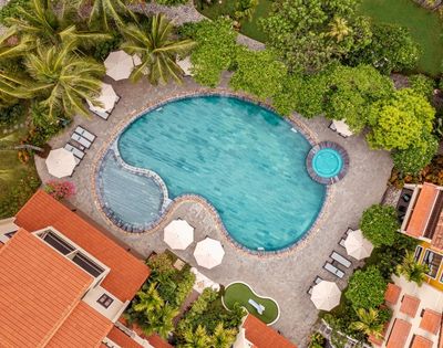 Aerial view of a resort pool shaped like a bean, surrounded by lush greenery and palm trees, with nearby lounge chairs and large umbrellas. Calm, inviting atmosphere.