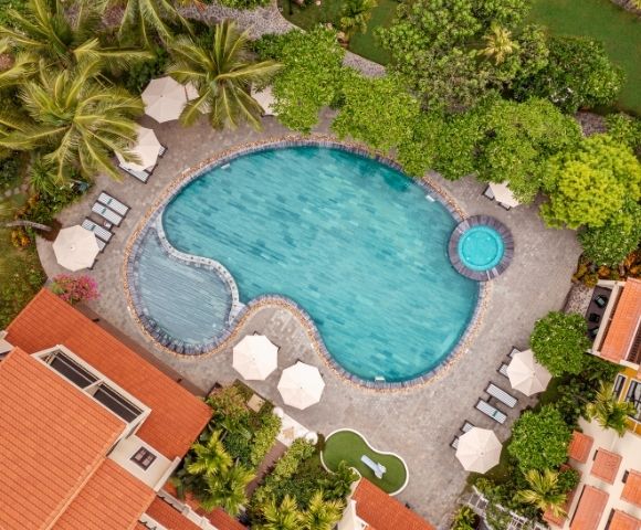 Aerial view of a resort pool shaped like a bean, surrounded by lush greenery and palm trees, with nearby lounge chairs and large umbrellas. Calm, inviting atmosphere.