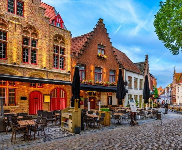 Cobblestone street with outdoor café seating, historic brick buildings with stepped gables, and vibrant evening lighting under a blue sky.