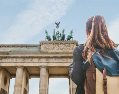 A woman with a backpack gazes at the impressive Brandenburg Gate in Berlin. The blue sky and historic monument convey a sense of awe and exploration.