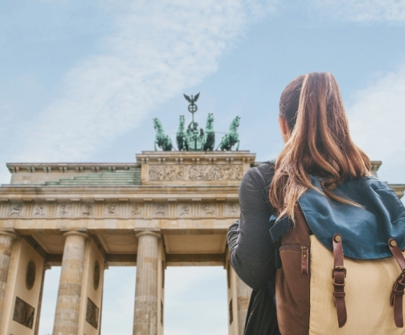 A woman with a backpack gazes at the impressive Brandenburg Gate in Berlin. The blue sky and historic monument convey a sense of awe and exploration.