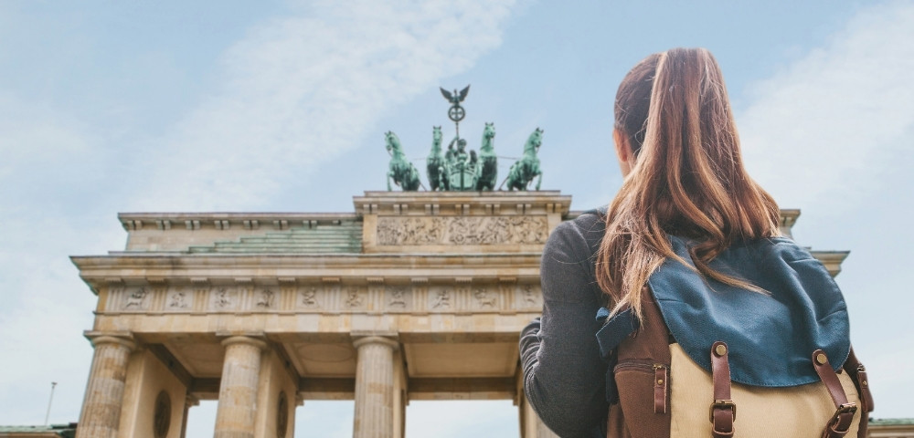 A woman with a backpack gazes at the impressive Brandenburg Gate in Berlin. The blue sky and historic monument convey a sense of awe and exploration.
