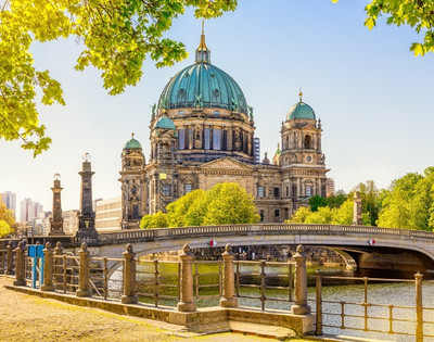 A grand cathedral with green domes sits beside a scenic river and stone bridge, surrounded by lush trees under a bright blue sky, evoking tranquility.