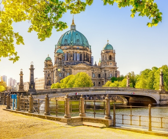 A grand cathedral with green domes sits beside a scenic river and stone bridge, surrounded by lush trees under a bright blue sky, evoking tranquility.