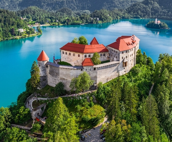 Aerial view of a historic castle with red roofs atop a forested hill, overlooking a serene turquoise lake surrounded by lush green mountains.