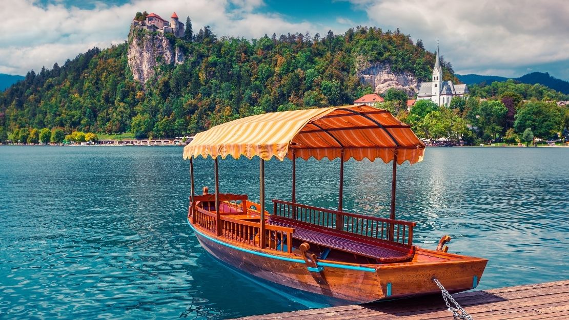 A traditional wooden boat with a yellow-striped canopy is moored on a serene lake. In the background, a green forested hill and a white church are visible.